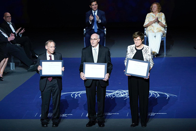 Robert Langer, Drew Weissman, and Katalin Karikó hold paper awards on a stage, while several people seated behind them clap