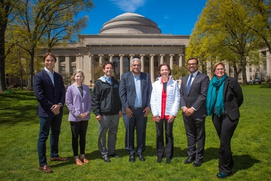 Photo of seven people posing on the grass in front of MIT’s Great Dome.