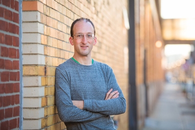 Photo of Michael Short leaning against an outdoor brick wall on a sunny day