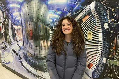 Photo of Tuba Balta posing beside a large image of the inside of a tokamak, a curved chamber with lots of shiny metal, knobs, and wires