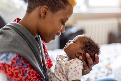 A young mother cradles her newborn child in her arms inside a white room with natural lighting.