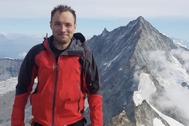 Photo of Octavian-Eugen Ganea, wearing a ski jacket and climbing ropes, standing high up on a mountain with another mountain peak behind him 