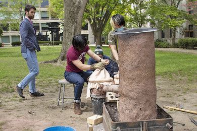 Photo of students working with a brown cylindrical furnace made from a mud-and-straw mixture that is nearly as tall as they are in a grassy courtyard