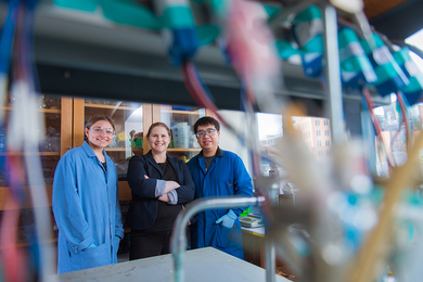 Photo of Ariel Furst, Rachel Ahlmark, and Gang Fan stand together in science lab, with many wires blurred in the foreground.