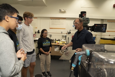 A group of four people stand in a 3D printing lab, listening to instruction from a lab manager, who is showing them a metal printed structure.