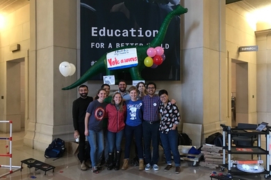 A group of nine people stands together, smiling. Behind them, above their heads, is an inflated dinosaur with a sign saying "Hello my name is Vote a saurus" on its back.