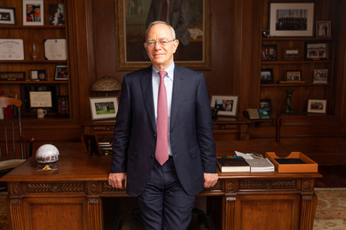 President L. Rafael Reif stands in his office, leaning slightly on his finely-detailed wooden desk. The wooden wall and bookshelves in the blurry background have oil paintings, photos, and mementos.