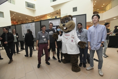 In the spacious, white atrium of MIT Building 46 filled with boards showing research posteers, four young men and a woman smile as they stand around MIT's Tim the Beaver mascot.