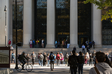 Photo of students crossing the street at 77 Mass. Ave., in front of MIT's main entrance, which features steps, large columns, and glass windows