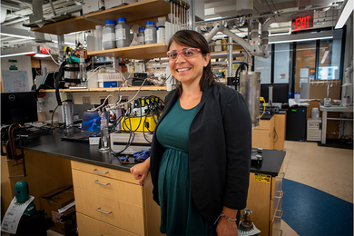Photo of Desiree Plata standing next to a lab bench stocked with lots of scientific and engineering equipment