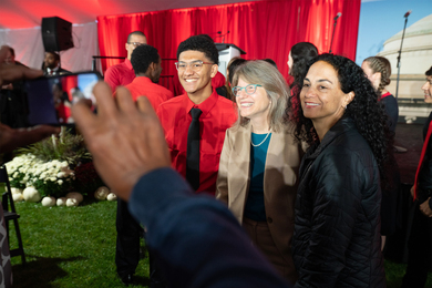 Sally Kornbluth, middle, poses for a picture with a member of The Choralaries in a red shirt, left, and another member of the MIT community in front of s red curtain