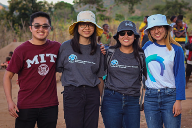 Four students wearing MIT T-shirts pose together with grasslands in the background. Half a dozen villagers of Mkutani, Tanzania, stand in the background.