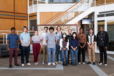 A row of 12 smiling students pose for a photo in the bright, modern lobby of MIT's Lincoln Laboratory. 