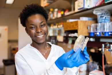 Rita Anoh smiles in a lab. She is wearing blue nitrile gloves and holding two petri dishes.