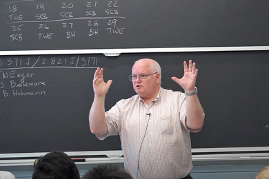 Photo of Tom Eagar standing and gesturing with both hands in the air in front of a classroom blackboard
