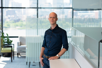 Photo of Nick van der Meulen standing next to an office cubicle, with an out-of-focus Charles River visible through the windows behind him..