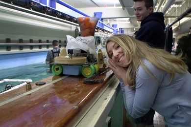 Woman leans on the ledge surrounding an indoor water tank, posing next to a small robotic boat that is also resting on the ledge