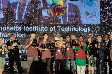 In a hail of confetti, the members of the MIT programming team stand on a large stage clutching trophies and plaques. Behind them, a huge screen displays the MIT team name.