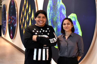 Alejandro Aguilera Castrejón and Melanie de Almeida stand together, in front of a long row of large, colorful scientific images