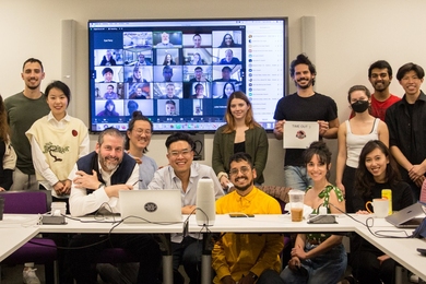 About a dozen people stand and sit around a table posing for a photo. Behind them is a screen with a five by five grid of people on a Zoom call