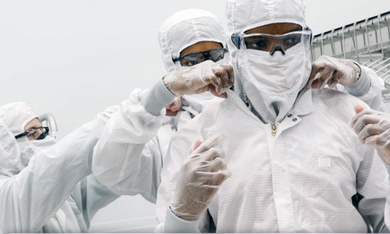 In a clean room, 3 people wear full-body clean suits. They help zip up each other’s suits.