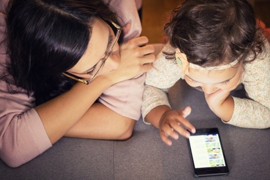 Overhead view of a mother observing her preschool-age daughter engaging with an educational app on a smartphone.