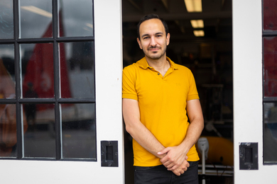 Fadel Adib stands between 2 open glass panel sliding doors while wearing a yellow shirt and black pants.