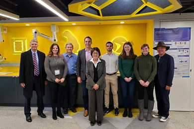 Photo of nine people posing in a line inside a colorful yellow-and-blue workshop