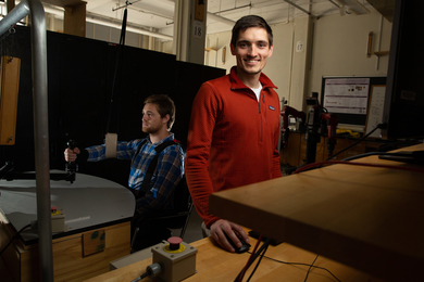 James Hermus stands at a raised desk in the lab and smiles at the camera. In the background, a person’s arm is strapped to a device and they are holding a joystick. 