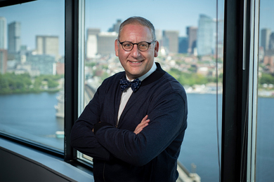Photo of Steven Spear in front of a wall of windows with the Charles River and Boston skyline in soft focus behind him.