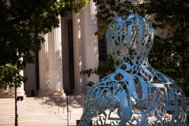 A view of the MIT campus. A sculpture of a human figure comprised of numbers and symbols is in the foreground.