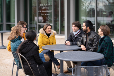 Esther Duflo sits with six students, all dressed for cold weather, at two outdoor sidewalk tables.
