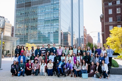 50 people pose in three rows in on the corner of a city block, with a large glass building dominating the background