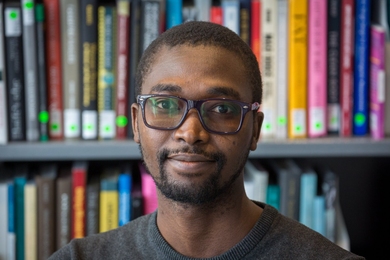 Head-and-shoulders photo of Abdullahi Tsanni standing in front of a bookcase filled with colorful books