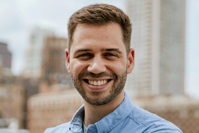 A bearded man wearing a button-down shirt with open collar smiles at the camera
