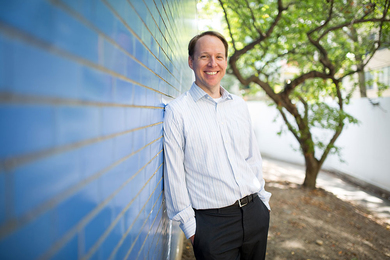 Benoit Forget leans against a blue tile wall outside, with an out-of-focus tree with green leaves in the background