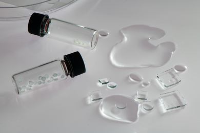 On a white table, 2 small glass vials contain pebble-like translucent chips. Some puddles of water are on the table, as well as circular and square chips of a hydrogel substance.