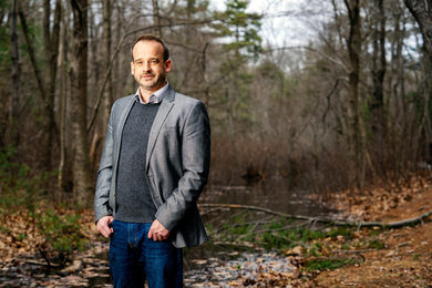 Fournier stands in the woods, and there is a stream in background. There are fallen leaves and branches, and it is a cloudy day in early Spring.