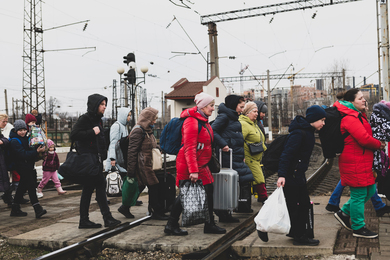 About 20 people of all ages, in winter coats and with lots of bags and luggage, cross train tracks on an overcast day.