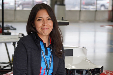 Cindy Heredia poses and smiles at the camera, sitting on her teams’ white, blue, and red driverless race car at the Indy Autonomous Challenge