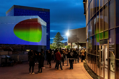 A crowd gathers at dusk between the Kresge Auditorium and MIT’s new Music Building, onto which an oval resembling a green-and-red heat map is being projected.