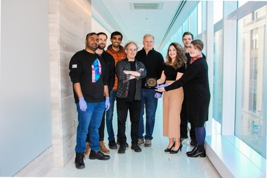 Eight people pose standing in a white hallway with the HUMANS nanowafer held between them. On their left is a wall, and the right are tall windows.