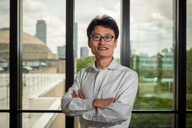 Jinhua Zhao stands with arms folded in front of a window with the Boston skyline in the distance