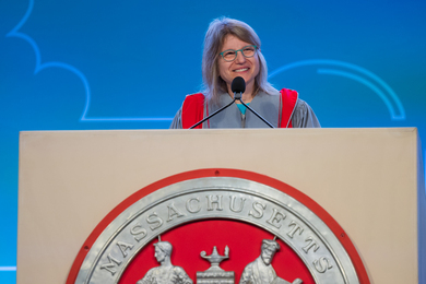 Sally Kornbluth, wearing grey and red academic regalia, speaks at a podium with a large MIT seal on the front.
