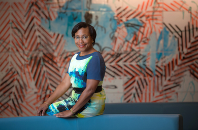 Paula Hammond poses for a portrait while sitting on the side of a couch. A mural with blue and orange is in blurry background.