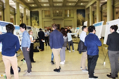 Roughly 20 students examine a large number of posters set up in a semicircle in an ornately decorated hall.