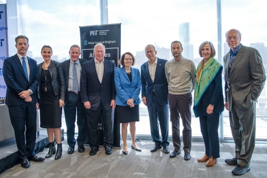 Nine academics pose standing side by side while smiling for the camera in a room with a glass wall and the Boston skyline behind them