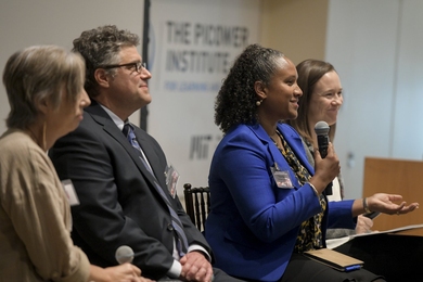 Sarah Milligan-Toffler, Gregory Bratman, Nsedu Witherspoon, and Sara Prescott sit on an auditorium stage. Witherspoon holds a microphone as she speaks to the audience out of frame.
