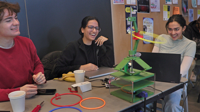 3 students smile at a table. A gyrating green structure as a small hula-hoop spinning around it.