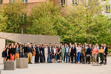 Photo of about 50 people standing in a line and posing outside of an MIT building made of brick and steel, with green trees behind them.